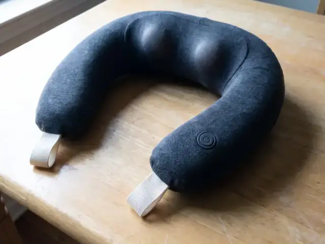 The generated product photo from the image-based page: a charcoal cordless neck-and-shoulder massager resting on a wooden table by a window.