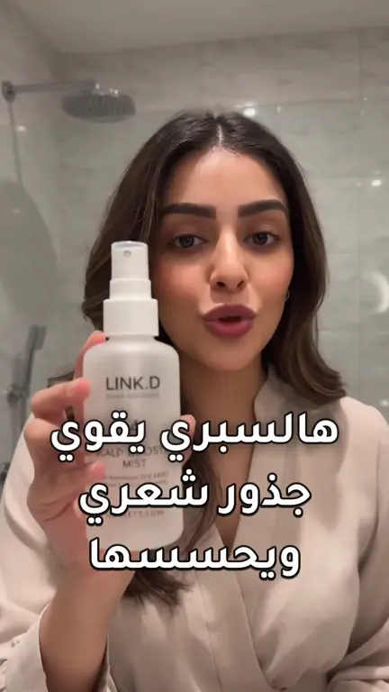First frame of the serum ad: a Gulf woman in a light beige blouse holding the frosted LINK.D Bond Defender bottle up to a phone camera in a bathroom, under the burned-in Arabic caption “هالسبري يقوي جذور شعري ويحسسها”.